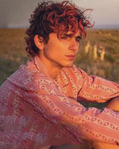 a young man with red hair sitting in the middle of an open field wearing a pink shirt