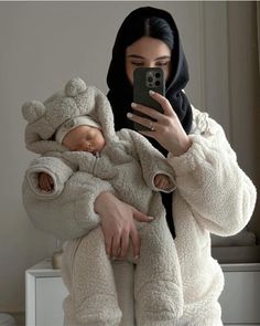 a woman holding a baby in a teddy bear outfit and taking a selfie with her cell phone