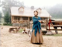 a woman standing next to a dog in front of a house with horses behind her