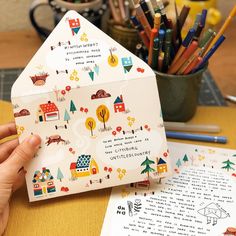 a person holding up a paper house with trees and houses on it, surrounded by colored pencils