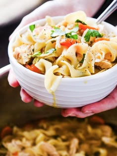 two hands holding a bowl of pasta with chicken and vegetables in it, while another person holds a spoon