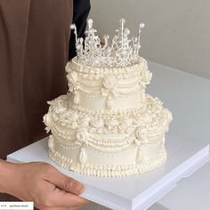 a person holding a white wedding cake on top of a table in front of the camera