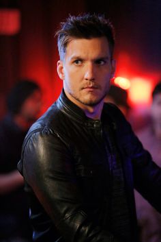 a man wearing a black leather jacket standing in front of a red light with his hands on his hips