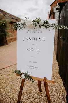 a wedding sign with greenery and flowers on it sitting in the middle of gravel