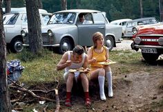 two people sitting on the ground eating food in front of old cars and parked vehicles