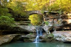 a small waterfall in the middle of a forest with a bridge over it's head