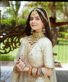 a woman in a white and gold bridal outfit holding a cell phone while standing under a tree