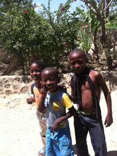 three young boys standing next to each other on a dirt ground with trees in the background