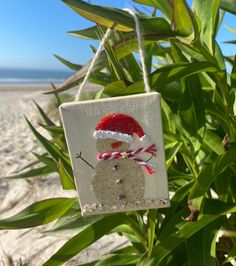 a snowman ornament hanging from a tree on the beach with green leaves