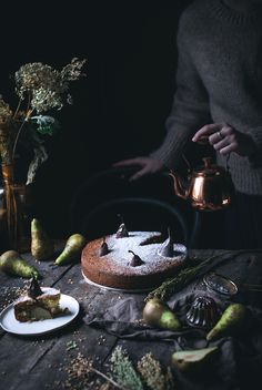 a cake on a table with pears around it and a teapot in the background