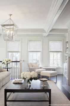 a living room filled with furniture and white flowers on top of a coffee table in front of two windows