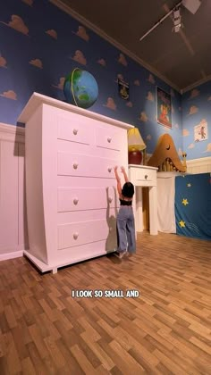 a little boy standing next to a dresser in a room with blue walls and clouds on the wall