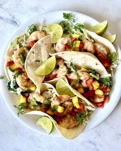 three shrimp tacos on a white plate with lime wedges