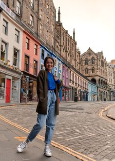 a woman standing in the middle of an empty street with buildings on both sides and people walking around