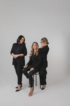 three women are posing for a photo in front of a white background, one is sitting on a chair and the other is standing