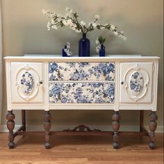 an ornate white and blue dresser with flowers on the top, in front of a gray wall