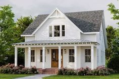 a white house with a porch and two windows on the first floor is surrounded by bushes and flowers