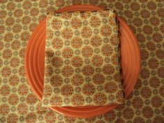 an orange plate with two napkins sitting on top of it next to a brown and white patterned table cloth