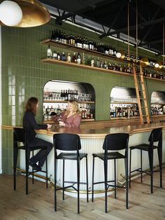 two women sitting at a bar with bottles on the shelves and stools in front of them