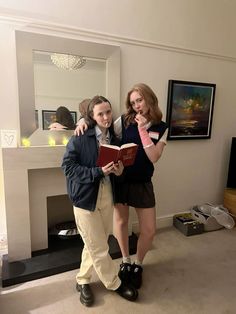 two people standing in front of a fire place holding a book and pointing at the camera