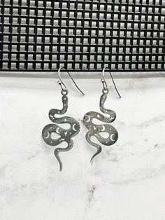 "Slither fashionably into the world with these stunning snake earrings. Made of stainless steel, they are durable and won't tarnish. Earring length is approx. 1 1/2\" long.  As with any piece of jewelry, it's best to take it off before showering, swimming, or participating in rough activities, such as sports." Silver Snake-shaped Metal Earrings, Silver Snake Shaped Metal Earrings, Silver Snake Shape Metal Earrings, Silver Snake-shaped Single Earring, Earrings Cottagecore, Cottagecore Earrings, Earrings Snake, Cottagecore Jewelry, Gemstone Earrings Dangle