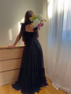 a woman in a long black dress standing next to a dresser with flowers on it