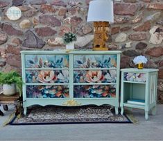 a blue dresser with flowers painted on it and a lamp next to it, in front of a stone wall