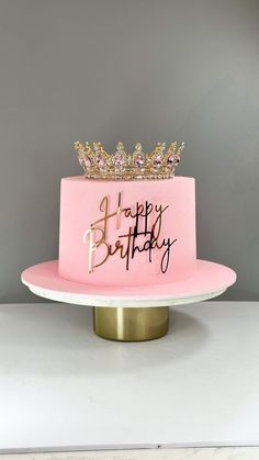 a pink birthday cake with gold crown and happy birthday written on the top, sitting on a white plate