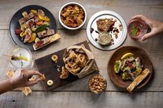 several plates and bowls of food on a wooden table with people holding wine glasses in front of them