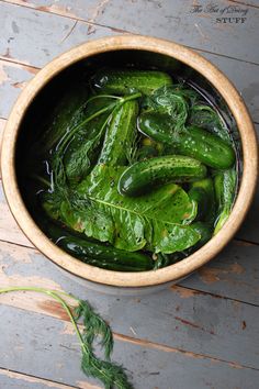 a wooden bowl filled with green pickles on top of a wooden table next to leaves