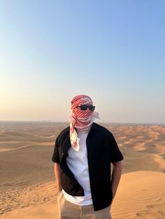 a man standing in the desert wearing sunglasses and a bandana on his head, looking at the camera