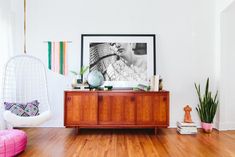 a living room with white walls and wood flooring is pictured in this image, there is a hanging chair next to the wooden cabinet