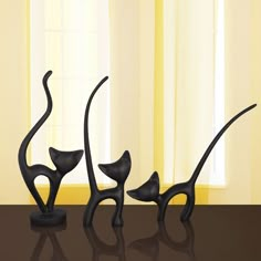 three black cat sculptures sitting on top of a table next to a window with yellow curtains