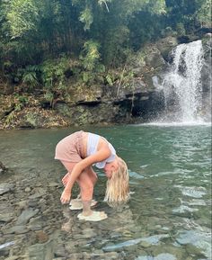 a woman bending over in the water next to a waterfall