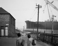 two people are walking down the street in front of a large ship that is being built