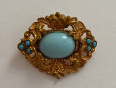 This beautiful Victorian brooch features a large blue cabochon stone, surrounded by smaller blue stones and mounted on gold metal.  The original C clamp is in working condition, and proves that this pin is over 100 years old.  A perfect gift for, or better yet something perfect to loan, the bride to be. If you like this posting, we ask you to please check out the other items in our shop.  *Note: Arioso Design strives to have a small footprint on our planet. Please know that your purchase may be Ornate Gold Cabochon Brooches, Blue Cabochon Brooches For Wedding, Blue Oval Brooch For Gifts, Antique Blue Jewelry With Brooch, Blue Cabochon Brooch For Formal Occasions, Formal Blue Cabochon Brooch, Blue Oval Brooch, Turquoise Cabochon Brooch As A Gift, Turquoise Cabochon Brooch For Gift