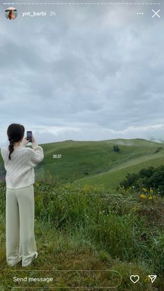 a woman standing on top of a lush green hillside holding a cell phone up to her face