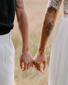 two people holding hands with tattoos on their arms and fingers, standing in a field