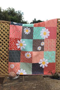 a colorful quilt with flowers on it sitting in front of a wicker fence next to a potted plant