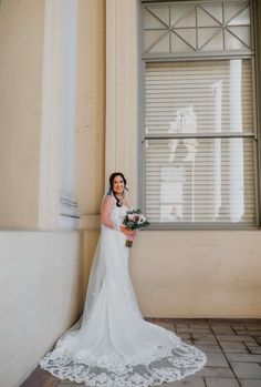 a woman standing in front of a window wearing a wedding dress and holding a bouquet