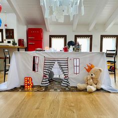 a teddy bear sitting on the floor in front of a table covered with a white cloth