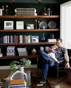 a man sitting in a chair holding a small child and looking at the camera with bookshelves behind him