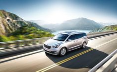 a silver minivan driving down a road next to the ocean with mountains in the background