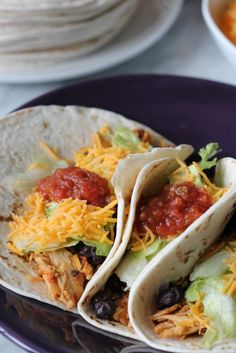 three tacos on a purple plate with cheese and salsa in the middle, along with tortillas