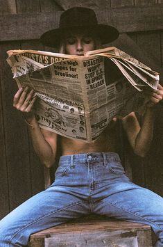 a woman sitting in a chair while reading a newspaper with her shirt off and hat on