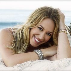 a beautiful woman laying on top of a sandy beach next to the ocean holding a pine cone