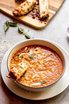 a bowl of tomato soup on a plate with some slices of pizza in the background