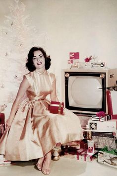 a woman sitting in front of a christmas tree with presents around her and a television