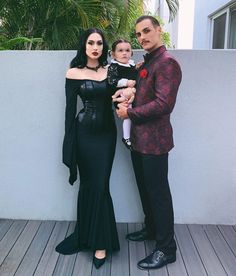 the man and woman are posing with their baby in front of a white wall outside