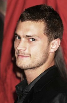 a close up of a person wearing a black suit and tie with a red curtain in the background